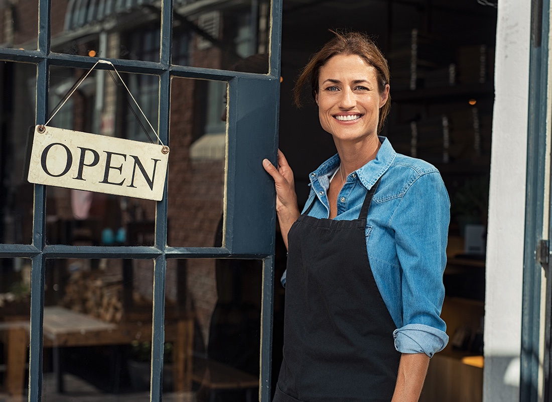 Business Insurance - Closeup Portrait of a Smiling Mature Business Owner Standing in the Front Door of Her Shop with an Open Sign