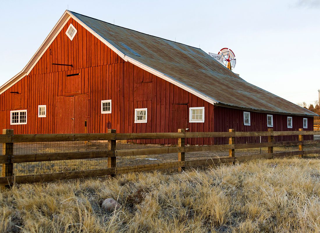 Insurance Solutions - View of an Old Red Barn Next to a Wooden Fence and Windmill on a Farm at Sunset