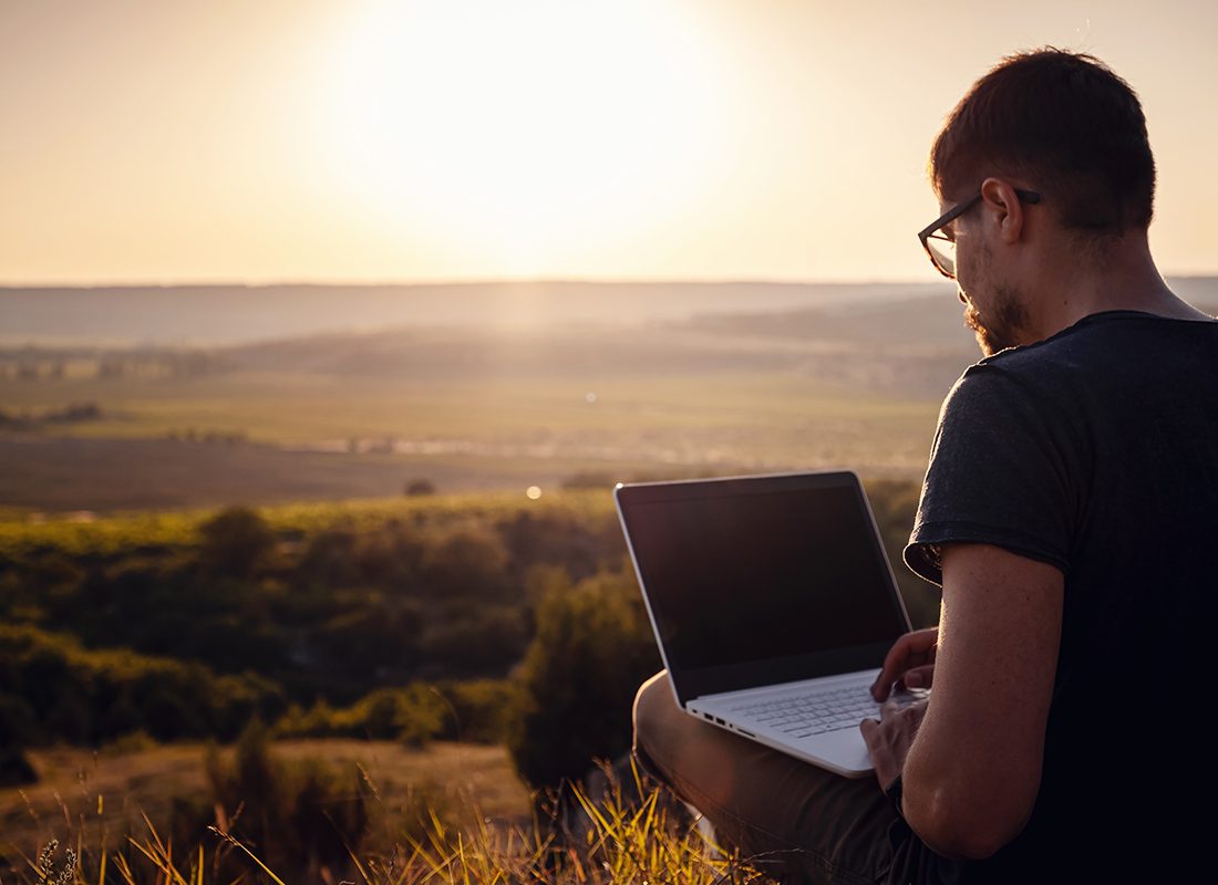Blog - Man Sitting in the Grass on His Laptop With Sun Shinning Over a Valley in Front of Him on a Beautiful Day